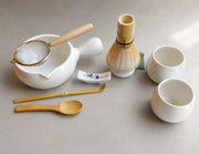 Matcha tool set with bamboo whisk, spouted ceramic bowl, mesh sifter, bamboo scoops, and white ceramic cups on a green tea mat