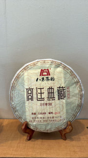 A round wrapped tea cake displayed on a small wooden stand against a plain light gray wall and wood surface. The wrapper is white with red and black Chinese text and decorative border details.
