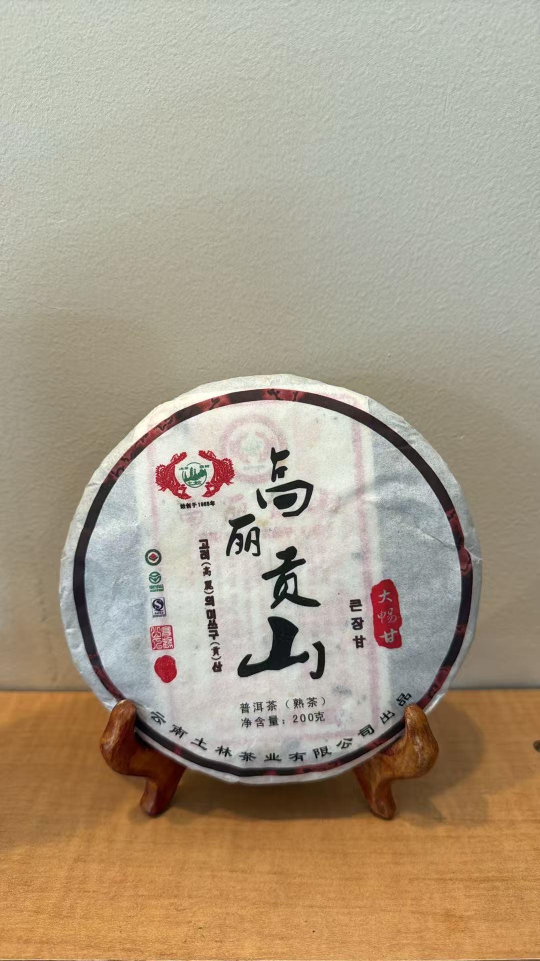 A round wrapped tea cake displayed on a small wooden stand against a plain wall, with the label showing Chinese characters and red branding details.