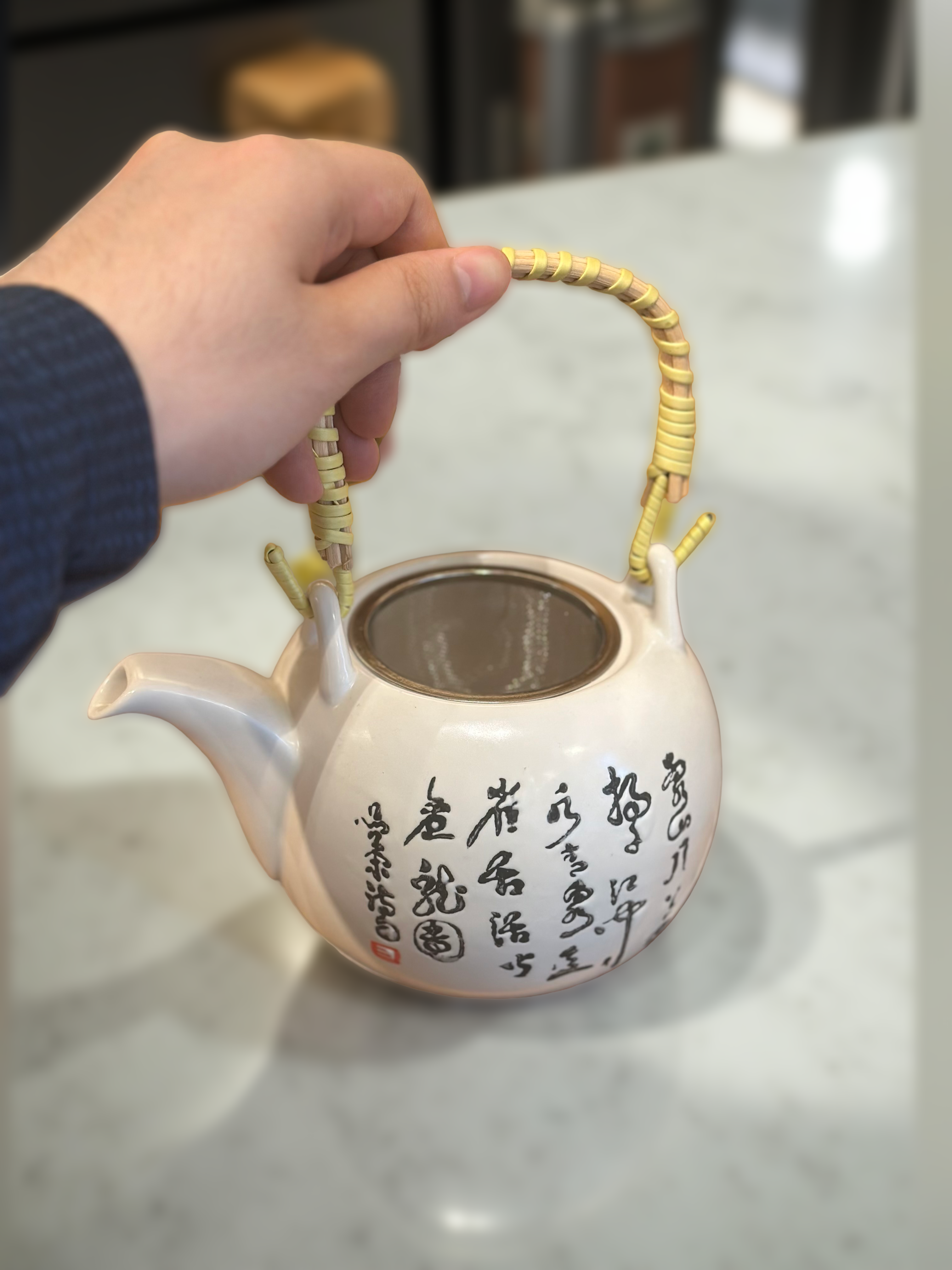 White teapot with black text and gold handle held by a hand on a light surface.