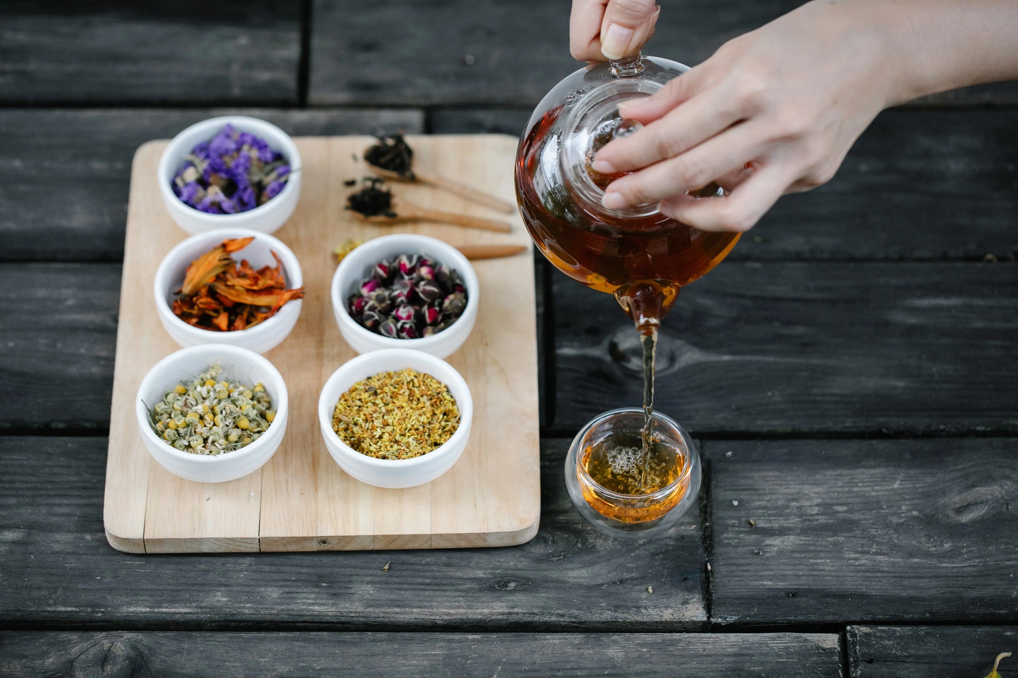 Person pouring liquid from a glass container into another on a wooden board with various dried herbs.