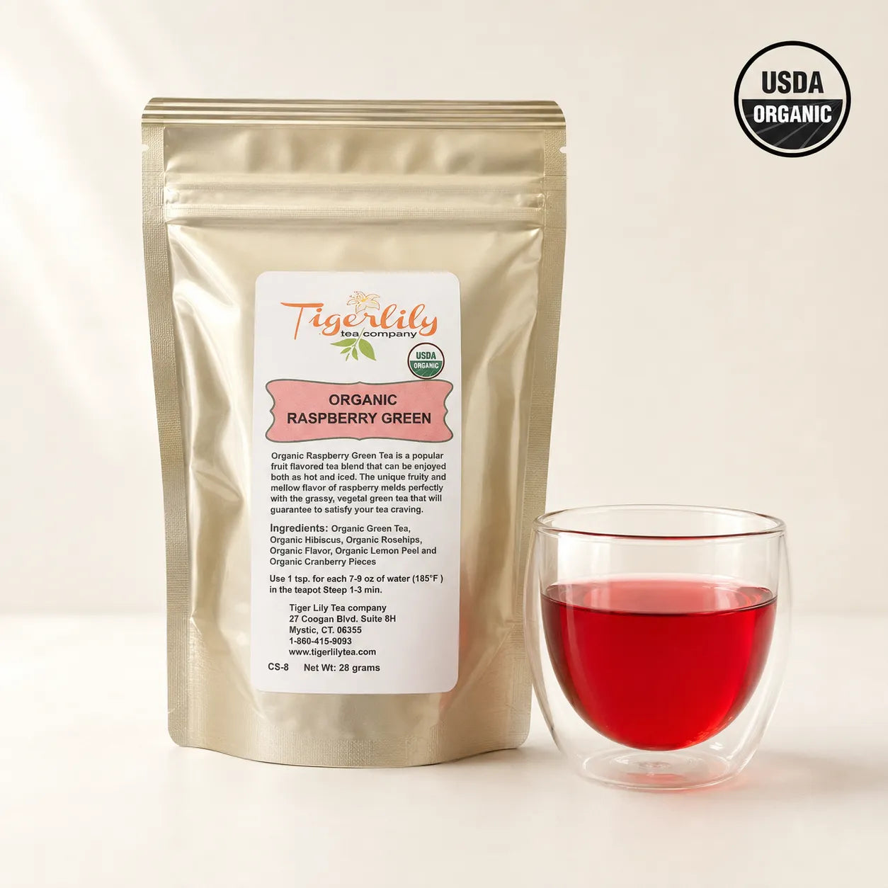 A stand-up pouch of Tigerlily Organic Raspberry Green tea next to a clear glass cup filled with the brewed tea, on a light beige background with a USDA Organic seal in the top right corner.